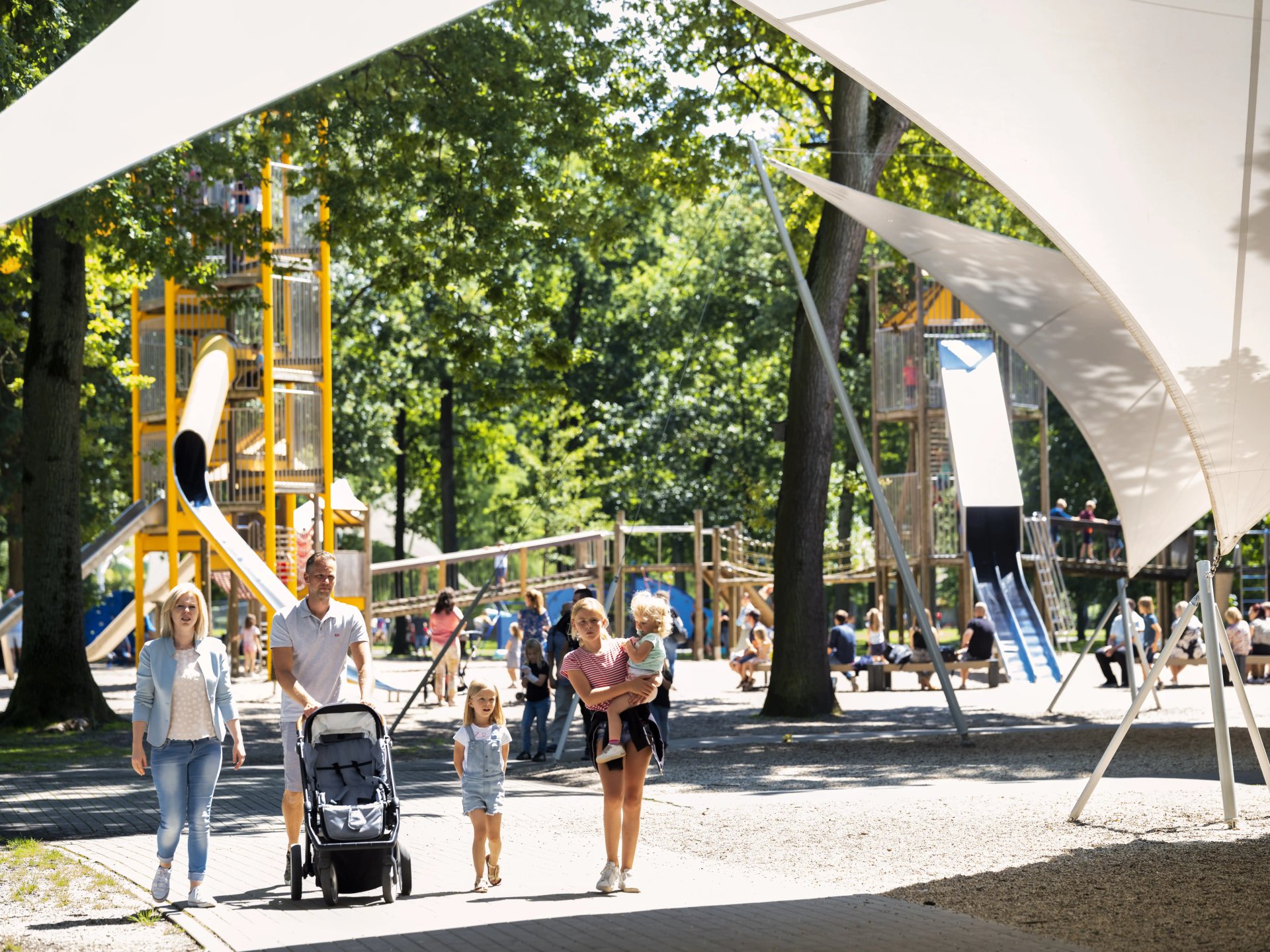 Gezinnen wandelen door de ruime speeltuin van Bokrijk met gele klimtorens en grote witte zonnezeilen tussen de bomen